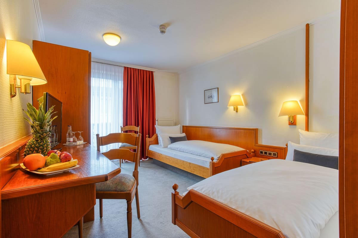 Ein Hotelzimmer mit einem Bett, einem Schreibtisch mit Obstschale, einem Sessel, einem Nachttisch und einem Fenster mit Vorhängen.