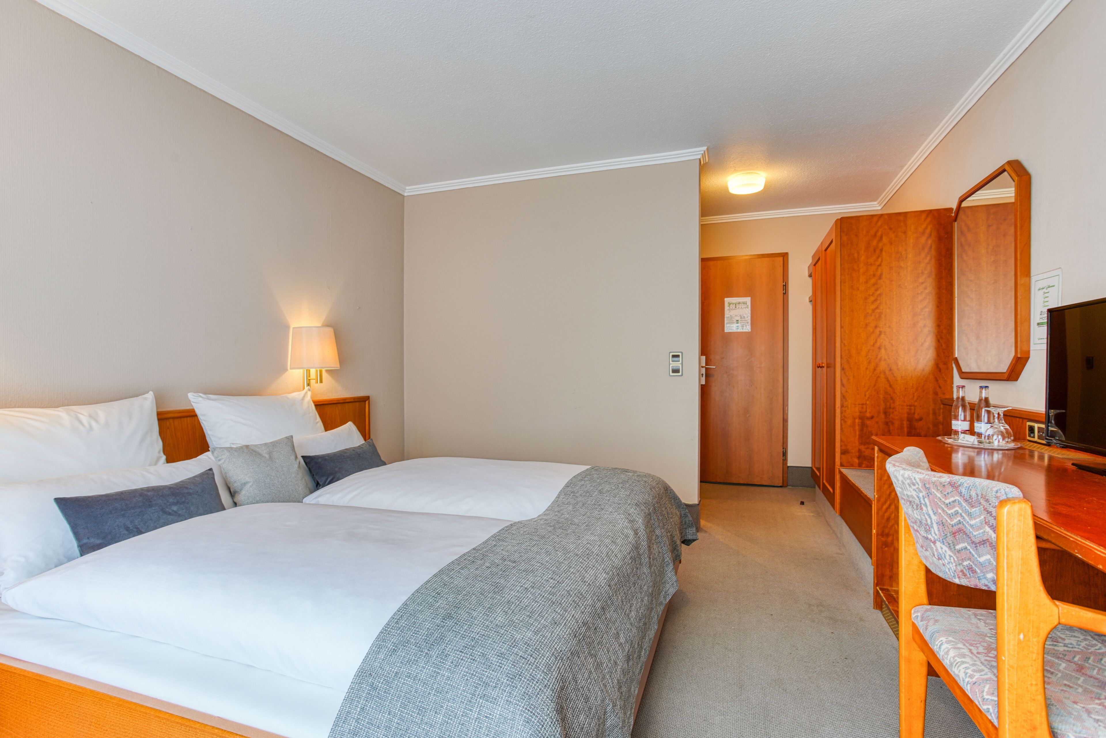 A hotel room with two beds, a desk, a chair, a lamp, and a television.