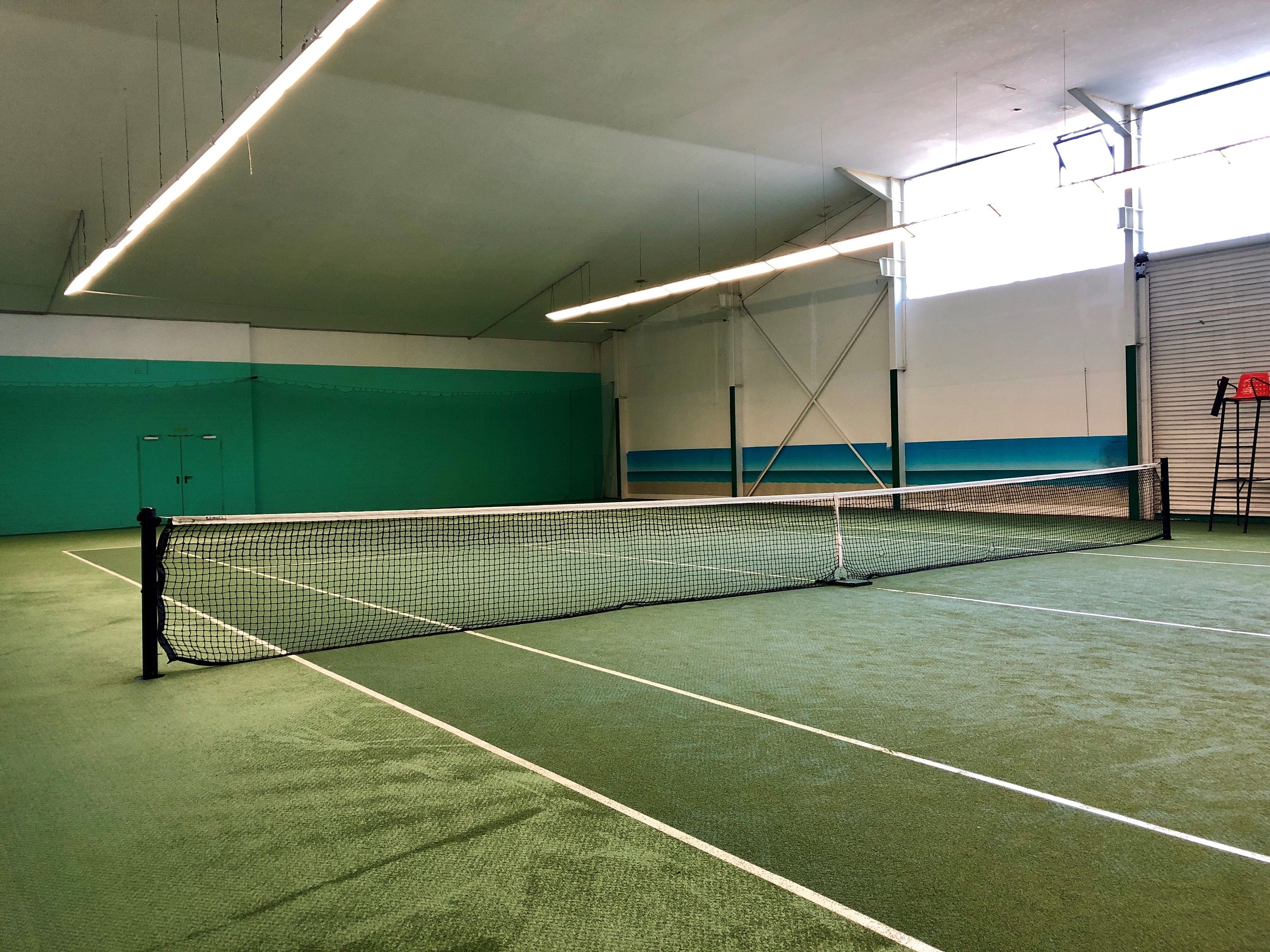 Una cancha de tenis cubierta con red, suelo verde y techo alto con iluminación fluorescente.