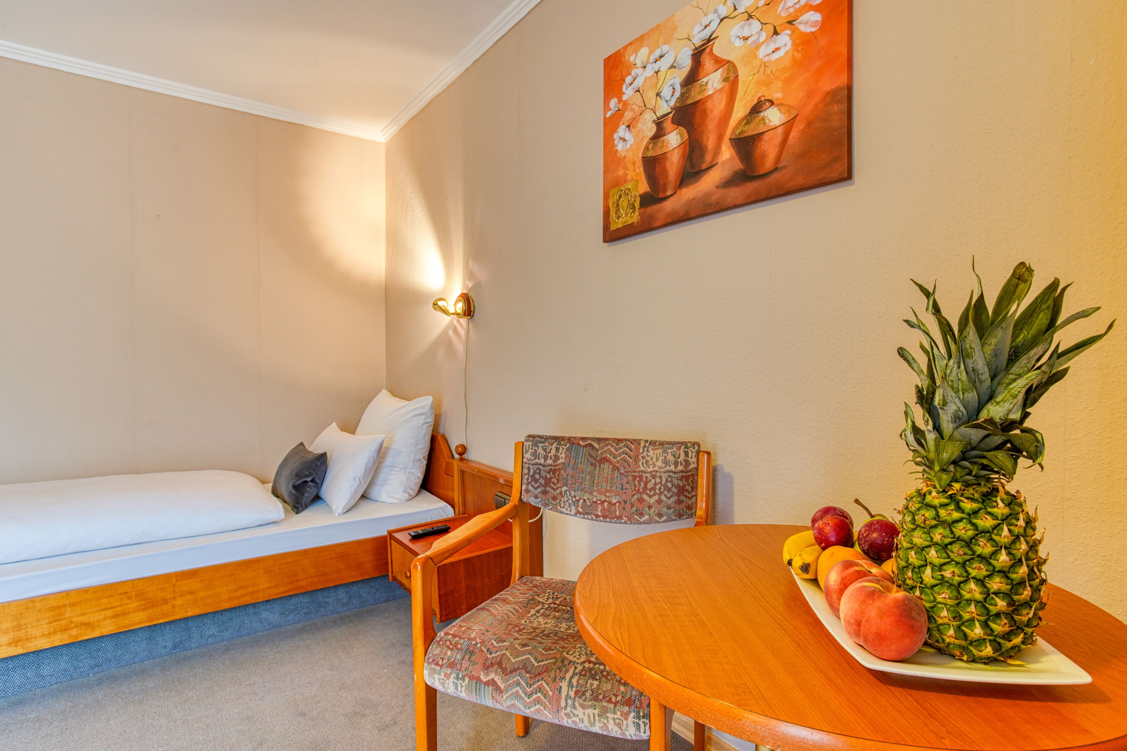 A compact room with a bed, a chair, a small table with fruit, and a painting on the wall.