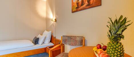 A compact room with a bed, a chair, a small table with fruit, and a painting on the wall.