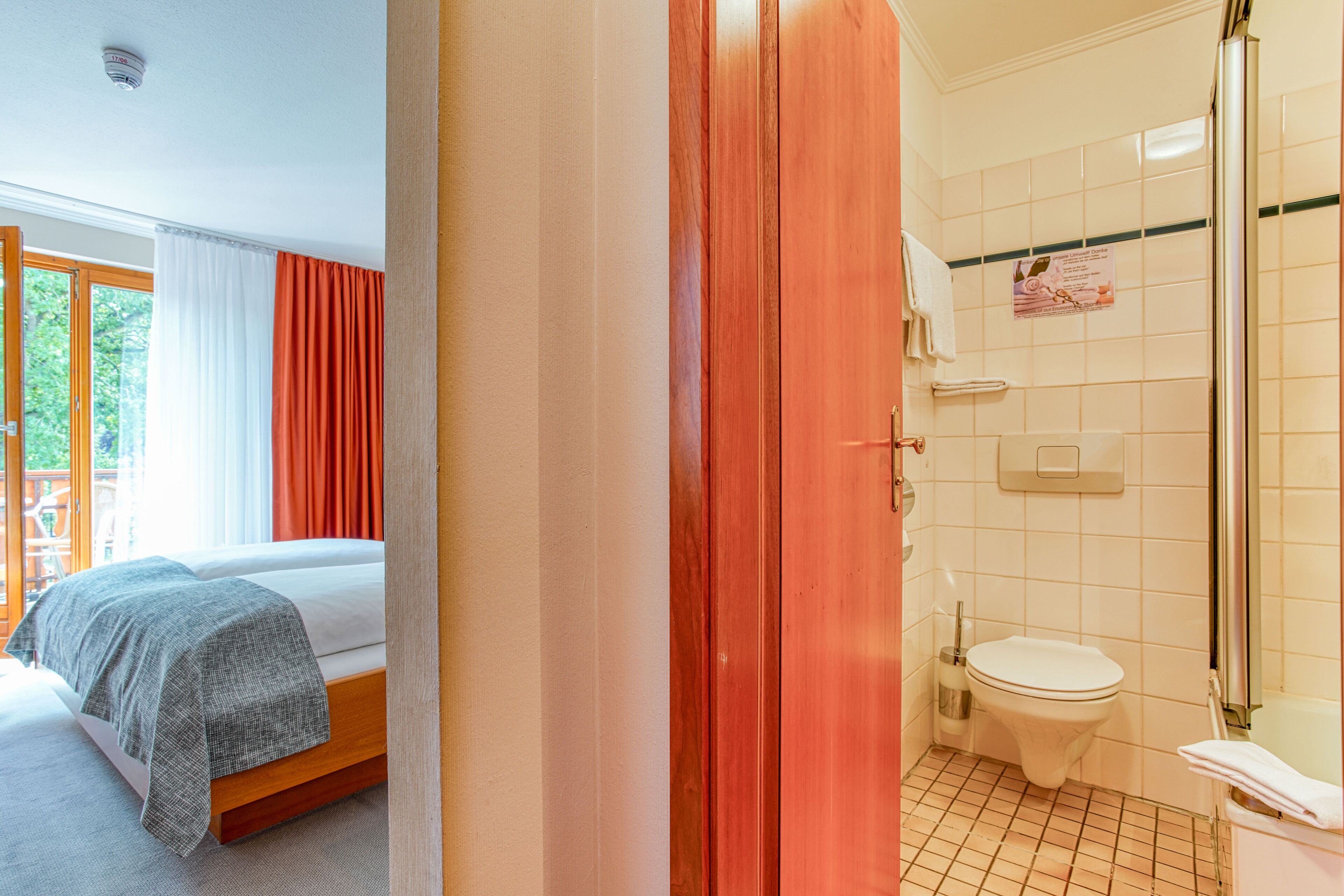 A hotel room with a bed, a bathroom with a toilet and shower, and a view of greenery outside.