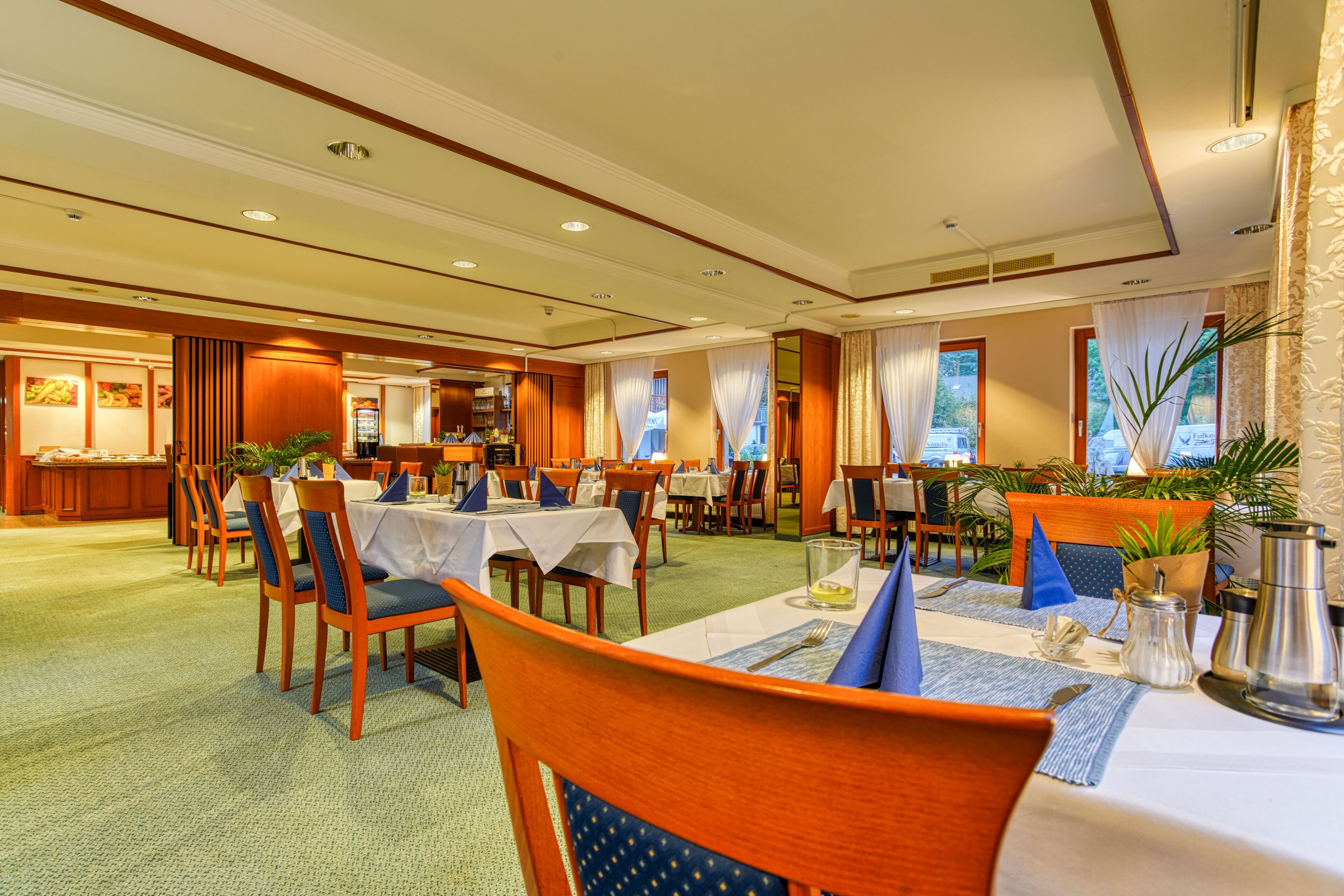 Amplia zona de comedor de hotel con varias mesas y sillas, una cocina al fondo y grandes ventanales con cortinas.