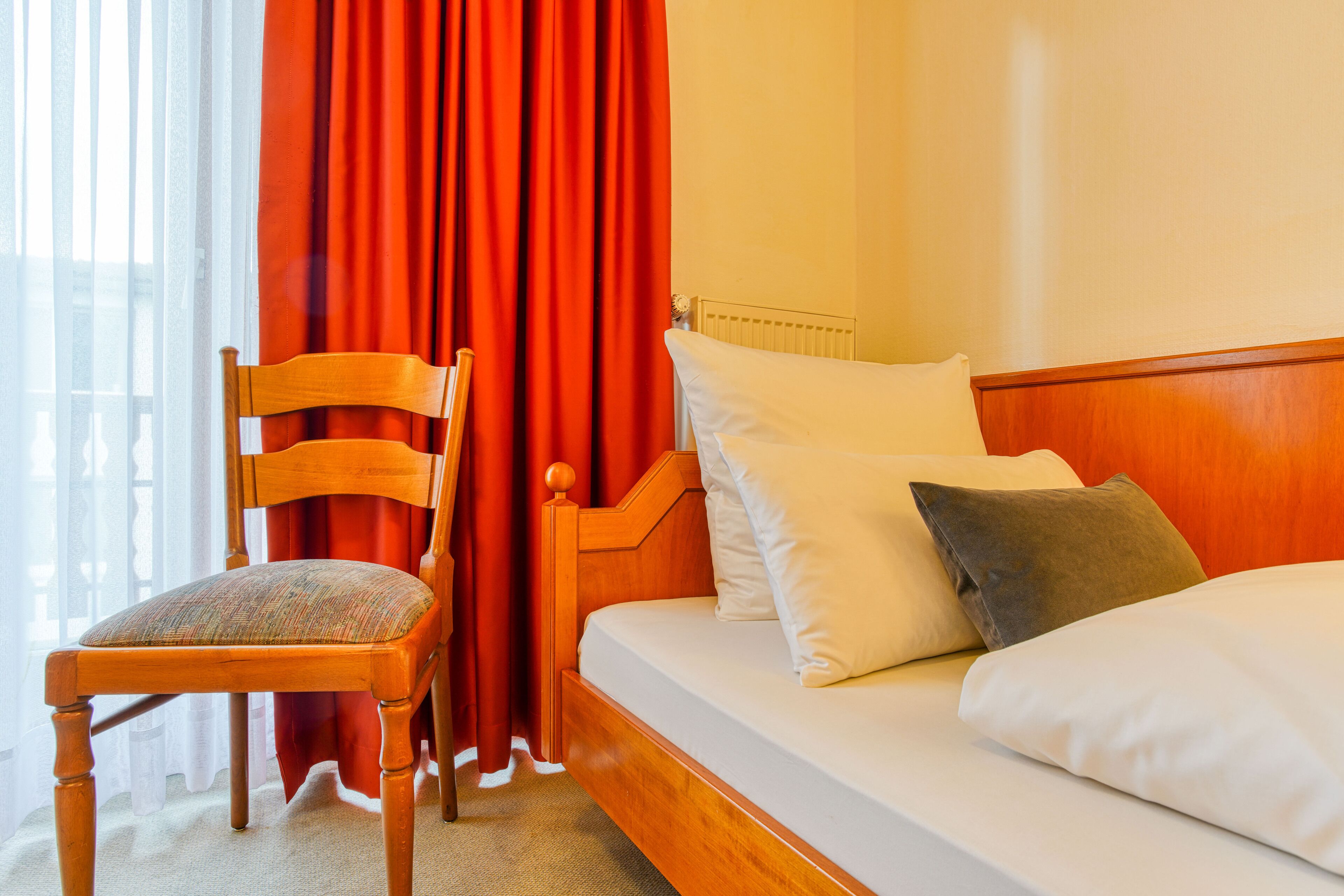 Una habitación de hotel con una cama y una silla de madera, y cortinas rojas.