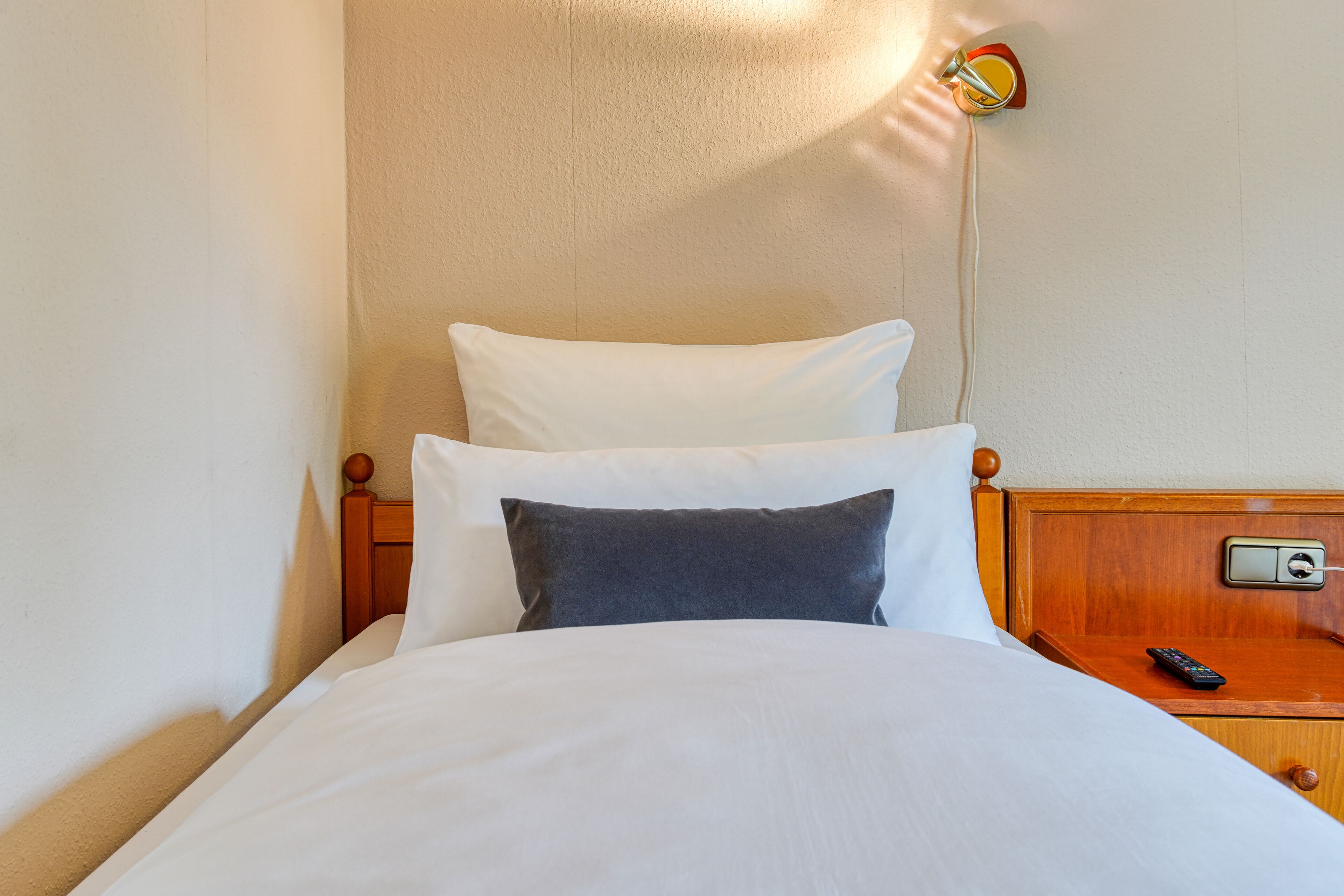 Ein ordentlich bezogenes Bett mit weißen und grauen Kissen, einem Holzkopfschild und einem Nachttisch mit Fernbedienung.