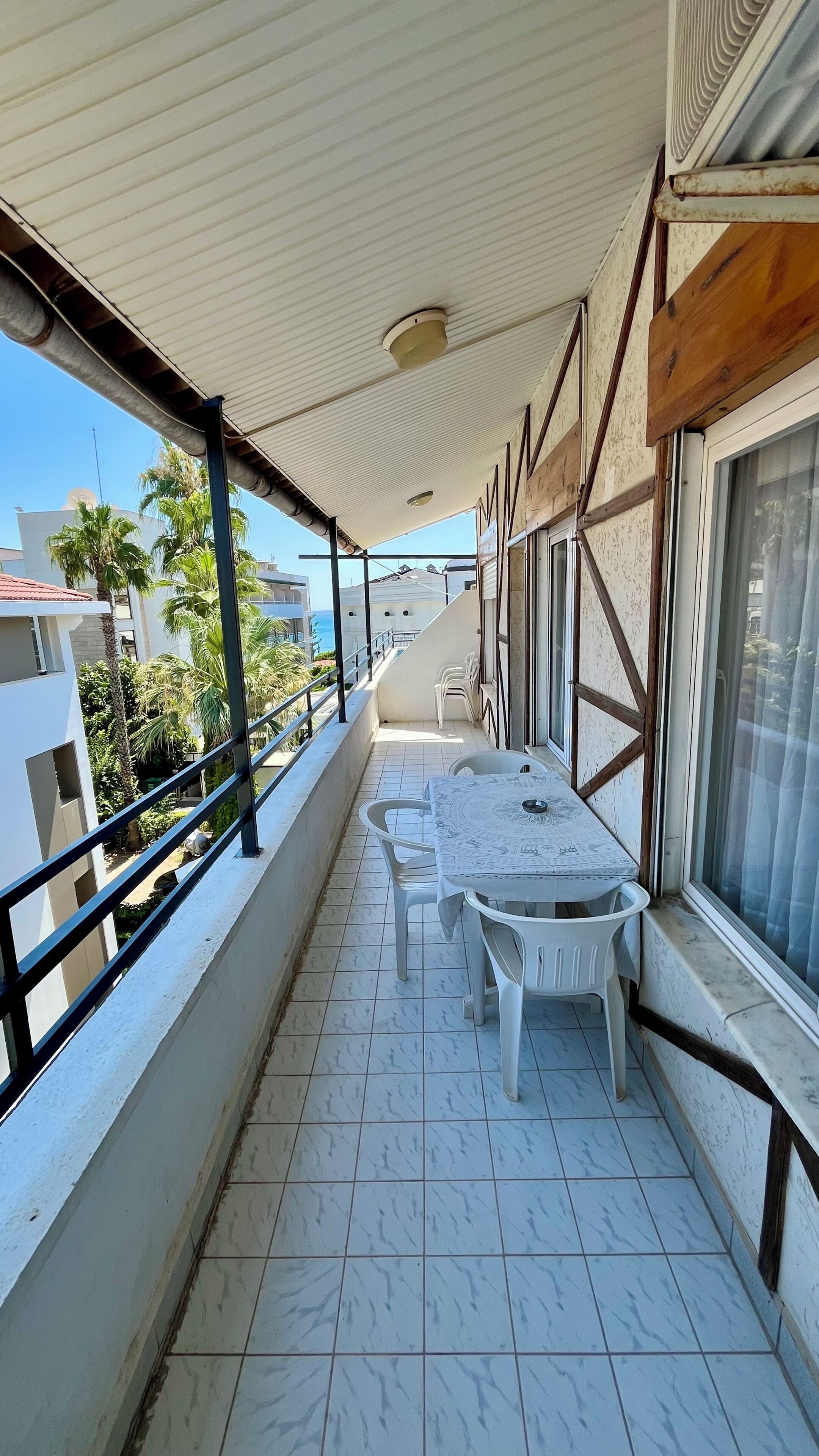 A covered balcony with a table and chairs, overlooking a residential area with palm trees and buildings.