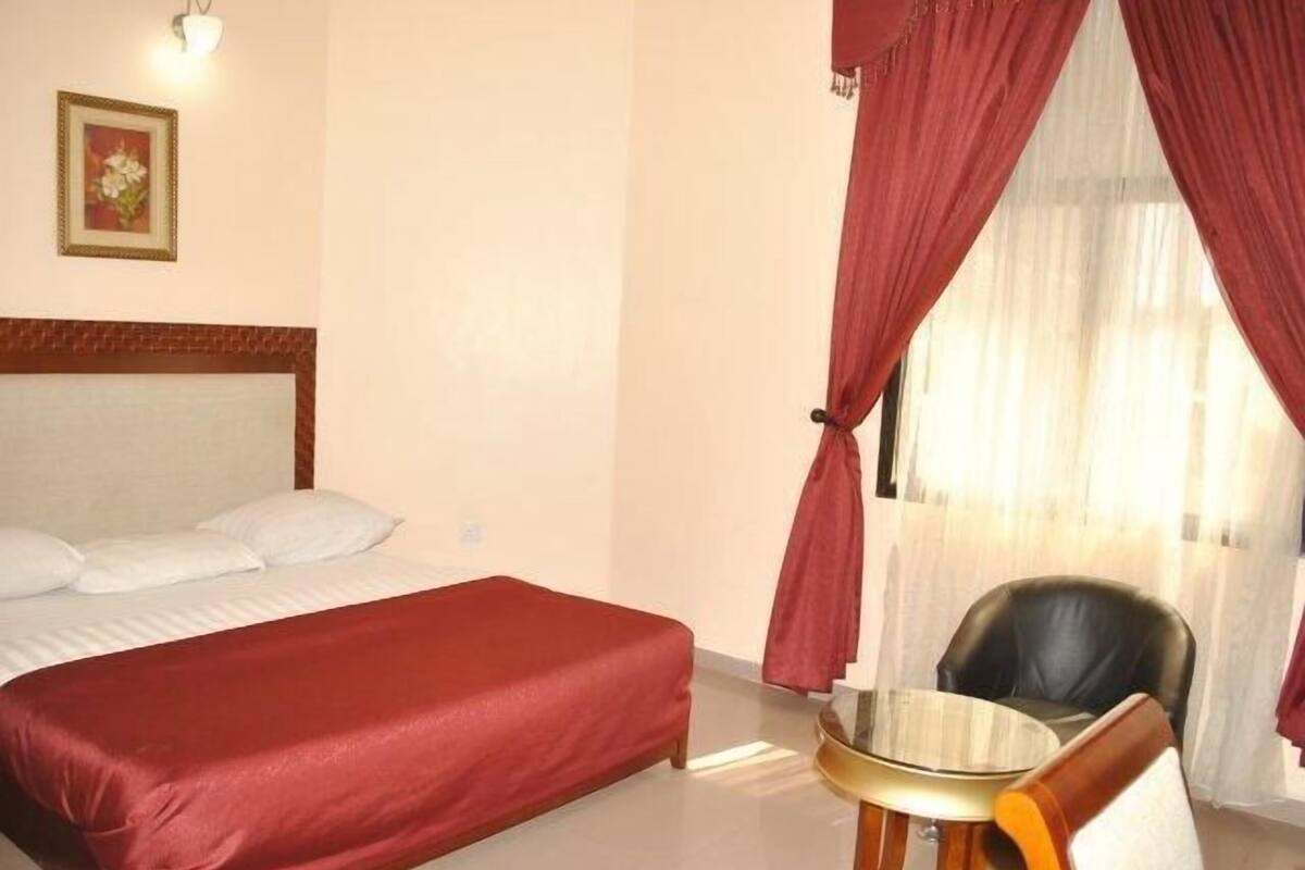 A hotel room with a bed, a small table, a chair, a picture on the wall, and red curtains.