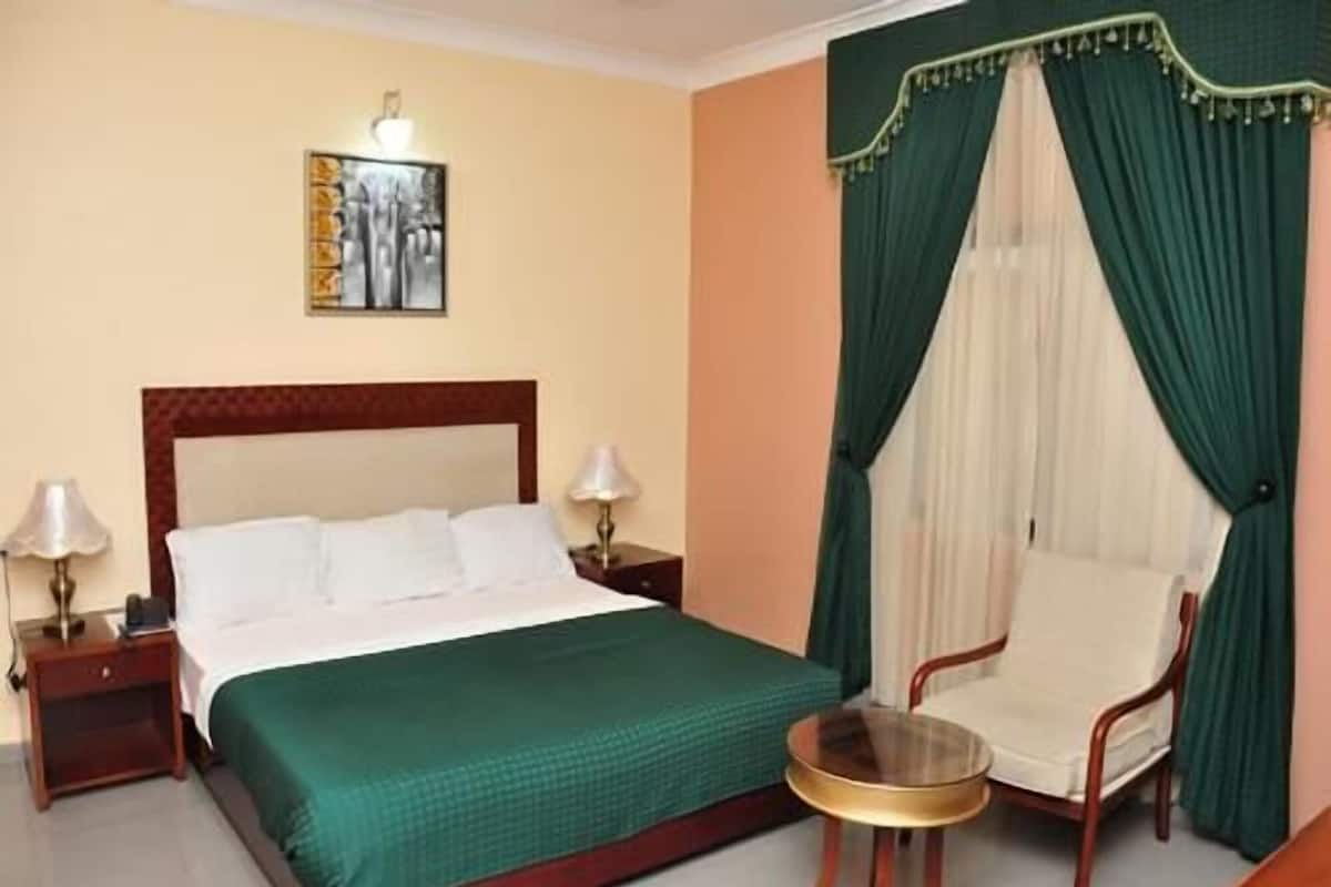 A hotel room with a bed, a chair, a small round table, a nightstand with a lamp, and a painting on the wall.