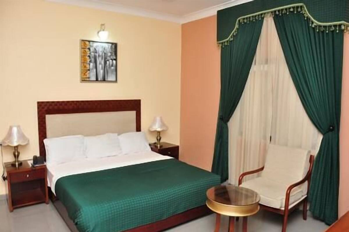 A hotel room with a bed, two bedside lamps, a chair, a small round table, and a painting on the wall.