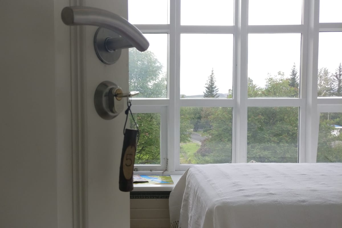 A room with a large window, a bed with a white cover, a door with a towel hanging on it, and a view of greenery outside.