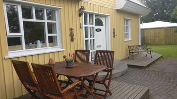 A yellow building with a white door and windows, a wooden deck with a table and chairs, and a small garden area.