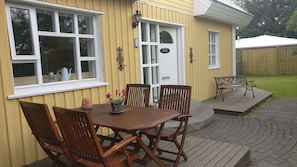 A yellow building with a white door and windows, a wooden deck with a table and chairs, and a small garden area. - Birta Guesthouse (Egilsstaðir)