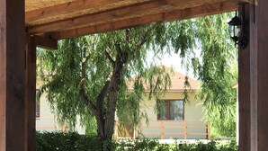A wooden pergola structure with a hanging lantern, a large tree with green foliage, and a building with a yellow facade in the background.