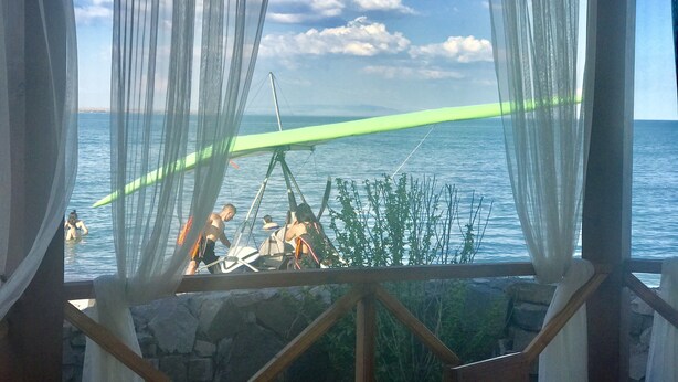 A view through sheer curtains of a calm sea with a green floating platform, a boat, and some greenery in the foreground.