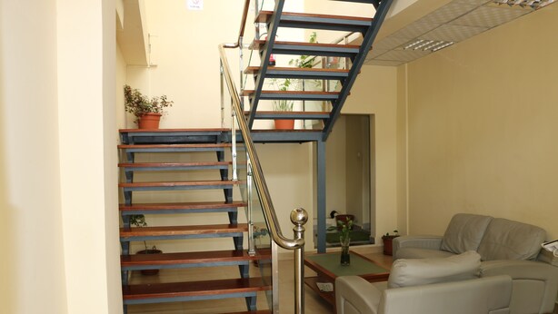 A staircase with wooden steps and metal railings, leading to an upper floor. There is a gray sofa and a small table with a vase in the living area. - SIS Hotel (Luanda)