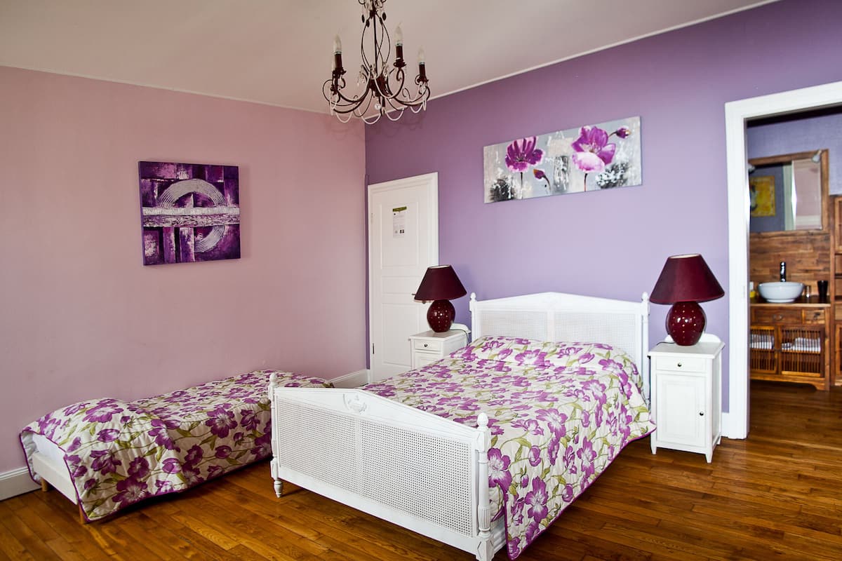 A bedroom with a double bed, two bedside tables, a nightstand, a dresser, a chandelier, and a door leading to another room.