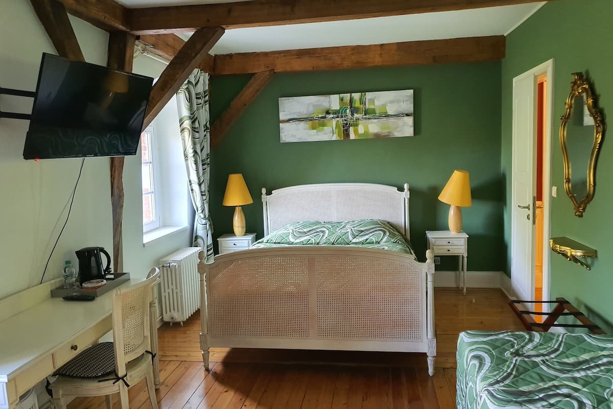 A bedroom with a bed, two bedside lamps, a television, a desk with a chair, a radiator, and a painting on the wall.