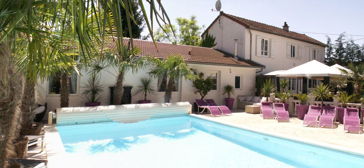 Une piscine entourée de palmiers et d’un bâtiment à toit de tuiles, avec des chaises longues et des parasols à proximité.
