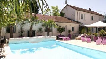 A swimming pool with lounge chairs and umbrellas, surrounded by palm trees and a building with a tiled roof.