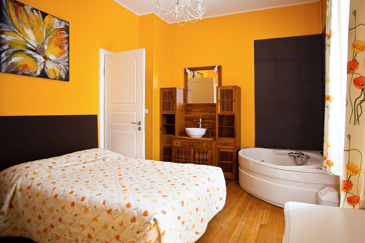 A bedroom with a bed, a wooden cabinet with a mirror, a white bathtub, and a bathroom with a shower curtain.