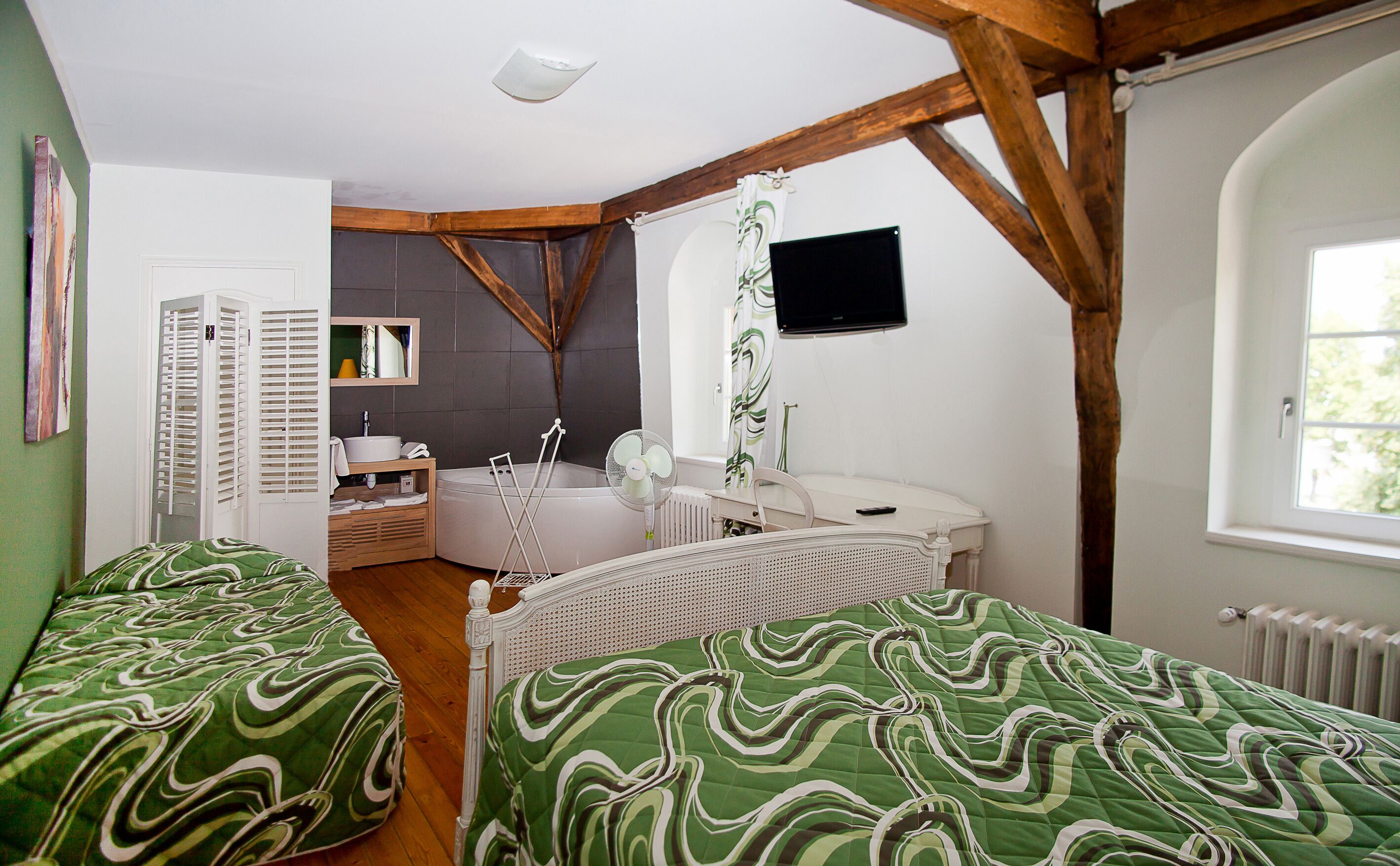 A bedroom with a bed, a bedside table, a mirror, a window, and a bathroom with a bathtub and a shower.