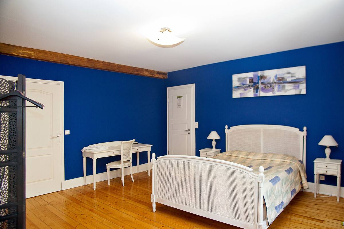 A bedroom with a white bed, a desk, a chair, a nightstand, and a wall-mounted lamp.