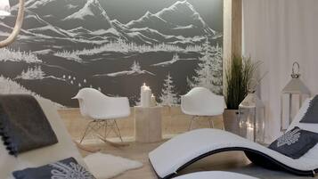 Un área de estar moderna con un mural de montañas, sillas blancas, una mesita con velas y una butaca con cojín.