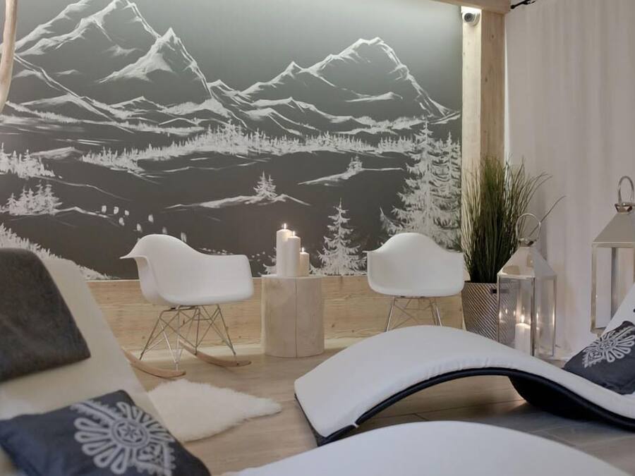 Un área de estar moderna con un mural de montañas, sillas blancas, una mesita con velas y una butaca con cojín.