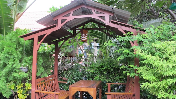 A wooden gazebo with two benches and a table, surrounded by lush greenery and potted plants.