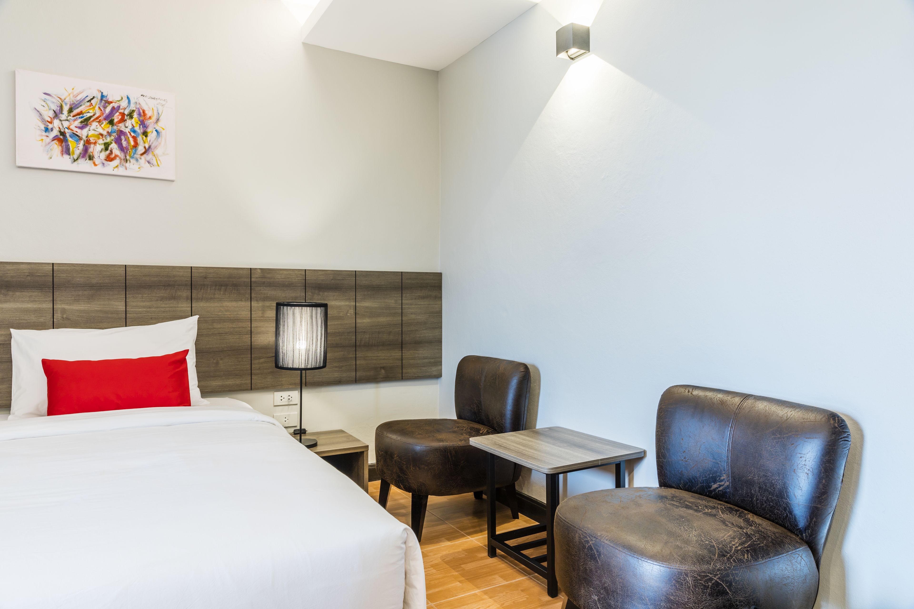 Habitación de hotel con cama, dos sillas, una mesita, una lámpara y un cuadro en la pared.