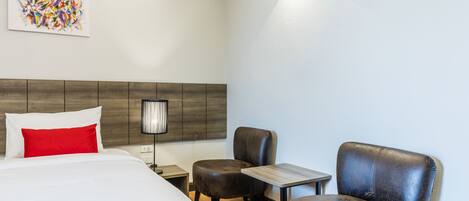 Habitación de hotel con cama, dos sillas, una mesita, una lámpara y un cuadro en la pared.
