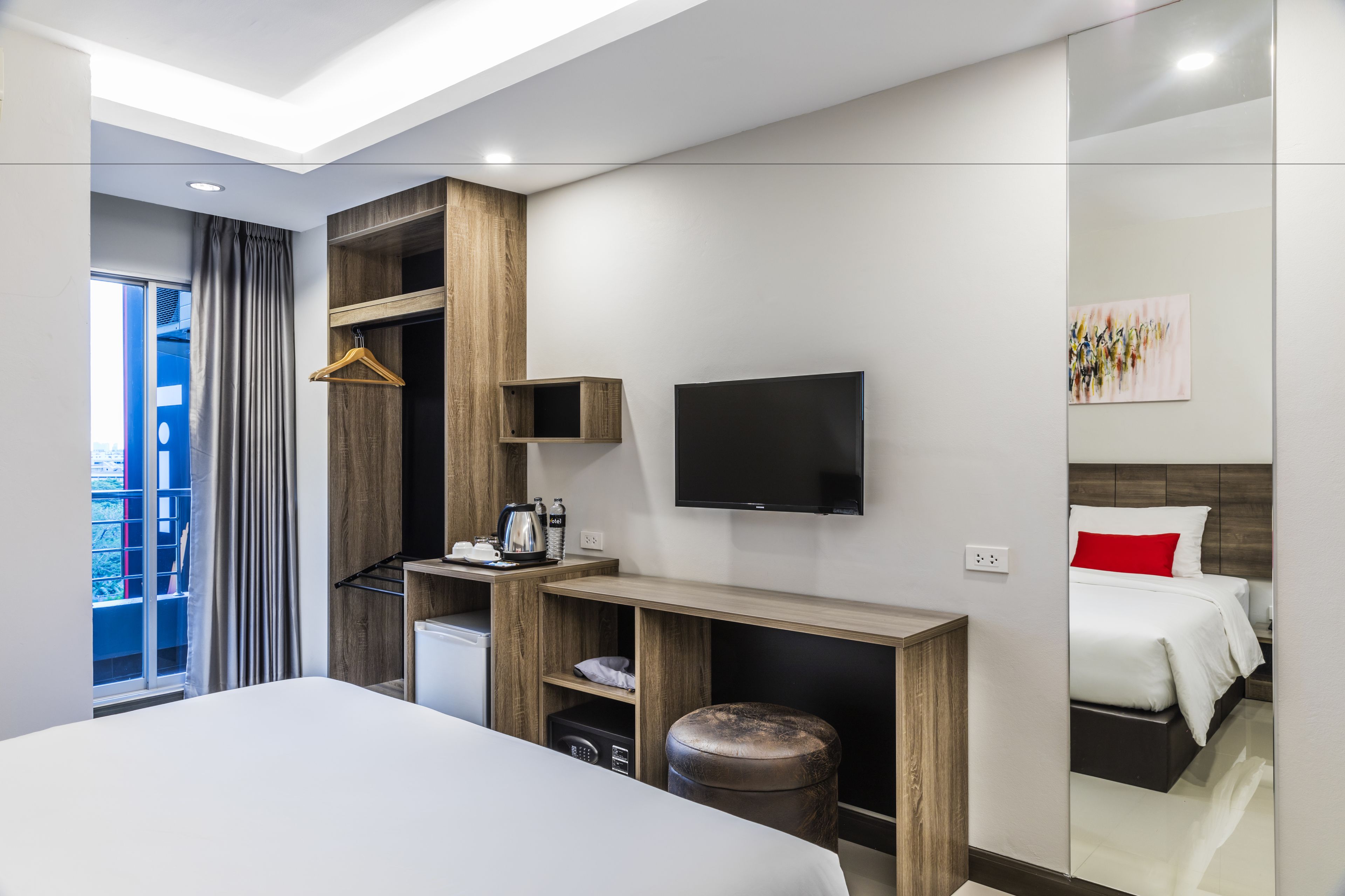 A modern hotel room with a large bed, a TV mounted on the wall, a wooden desk, and a small kitchenette.