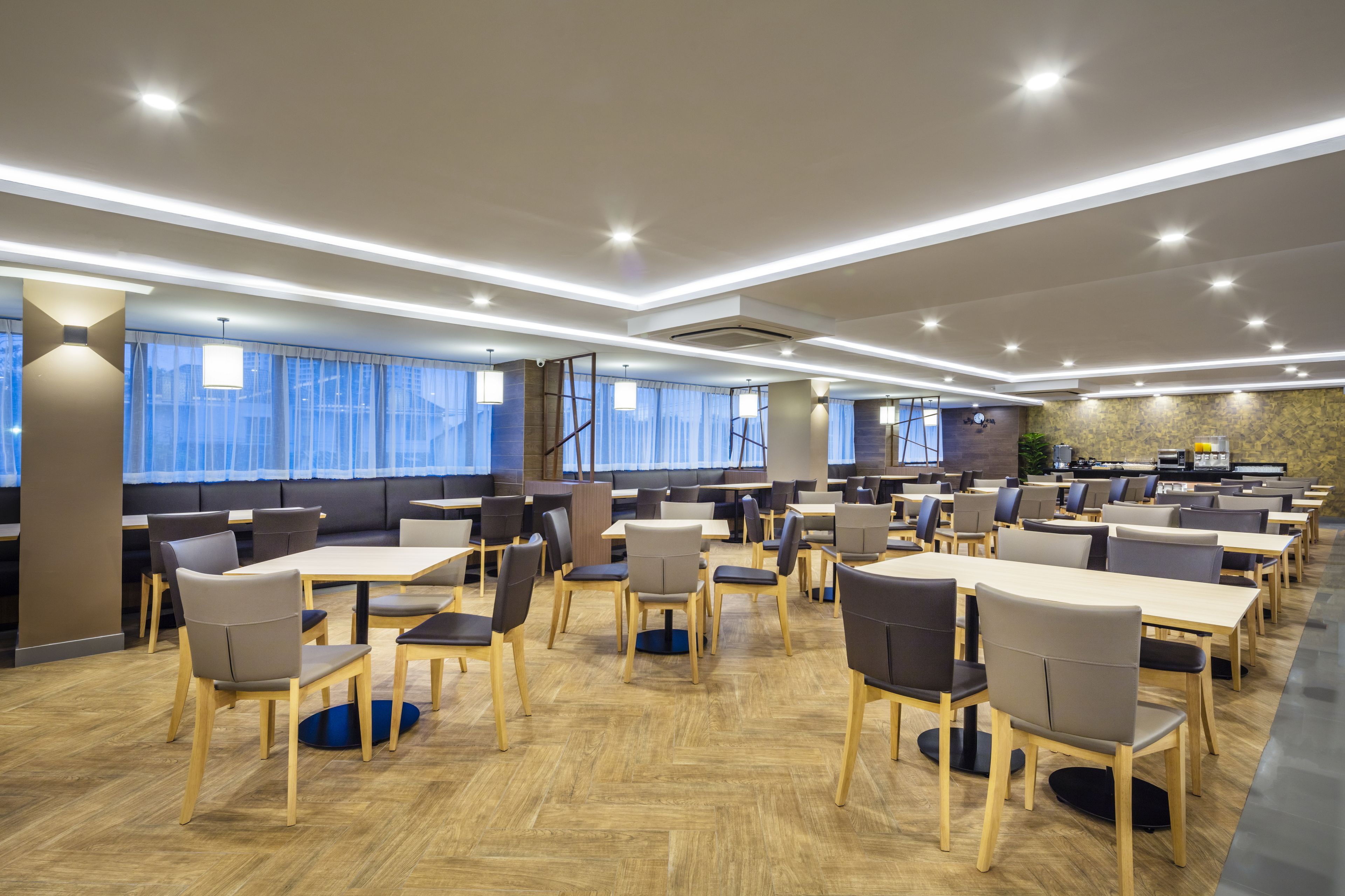A spacious dining area with multiple tables and chairs, large windows with curtains, and a modern ceiling design.