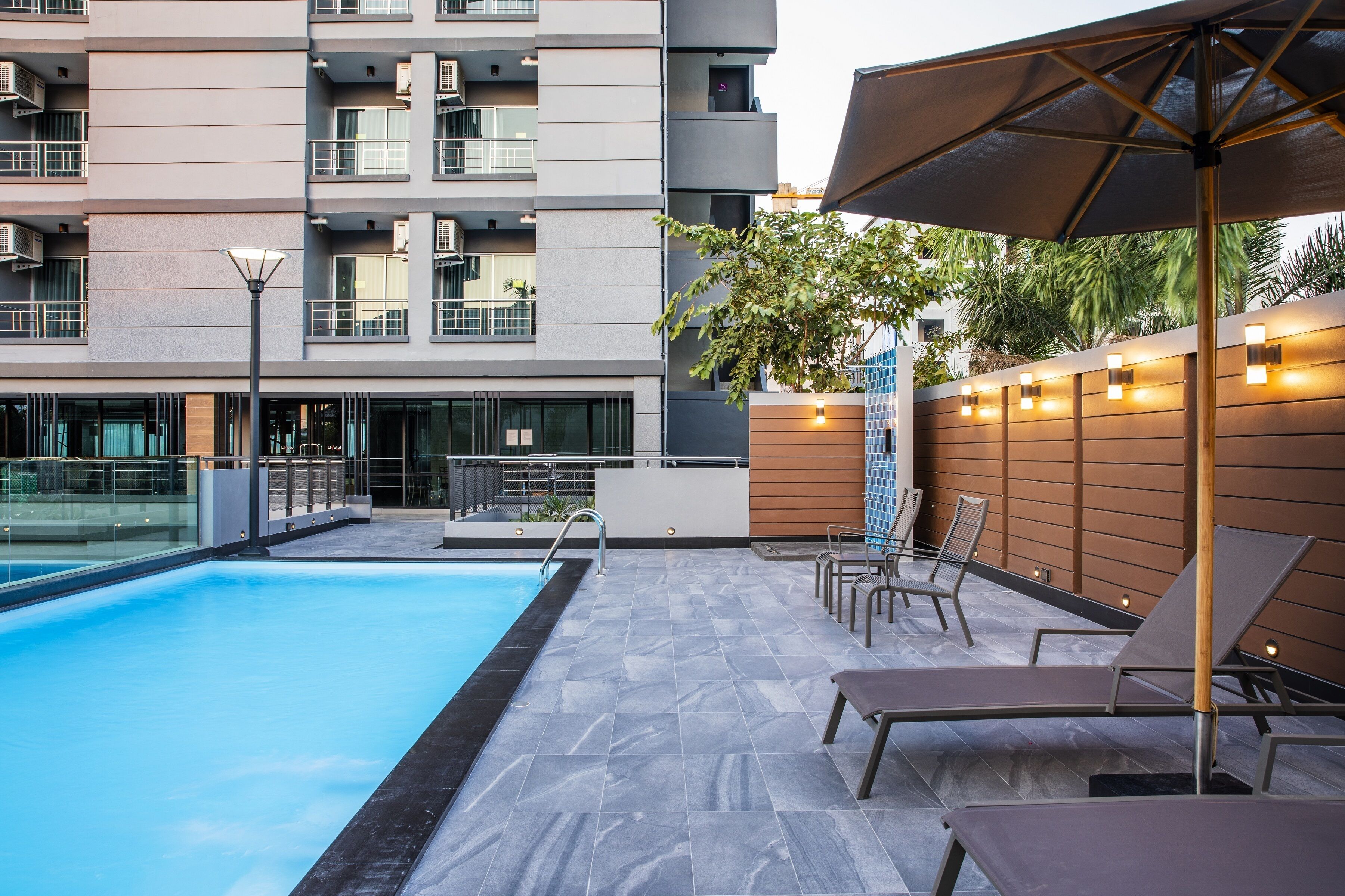 A swimming pool with lounge chairs and an umbrella, surrounded by a modern building with balconies and outdoor lighting.