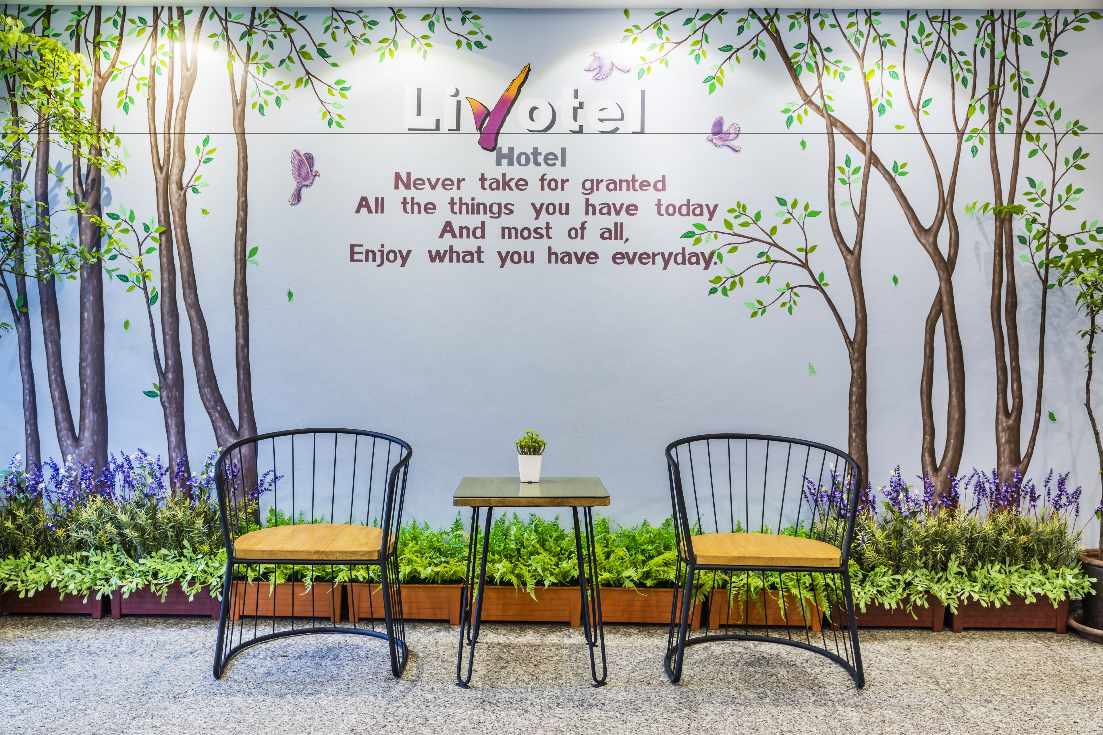 Une pièce avec deux chaises et une petite table, un mur orné d’une fresque représentant un arbre et d’une citation inspirante, ainsi que des plantes en pot disposées autour des chaises.