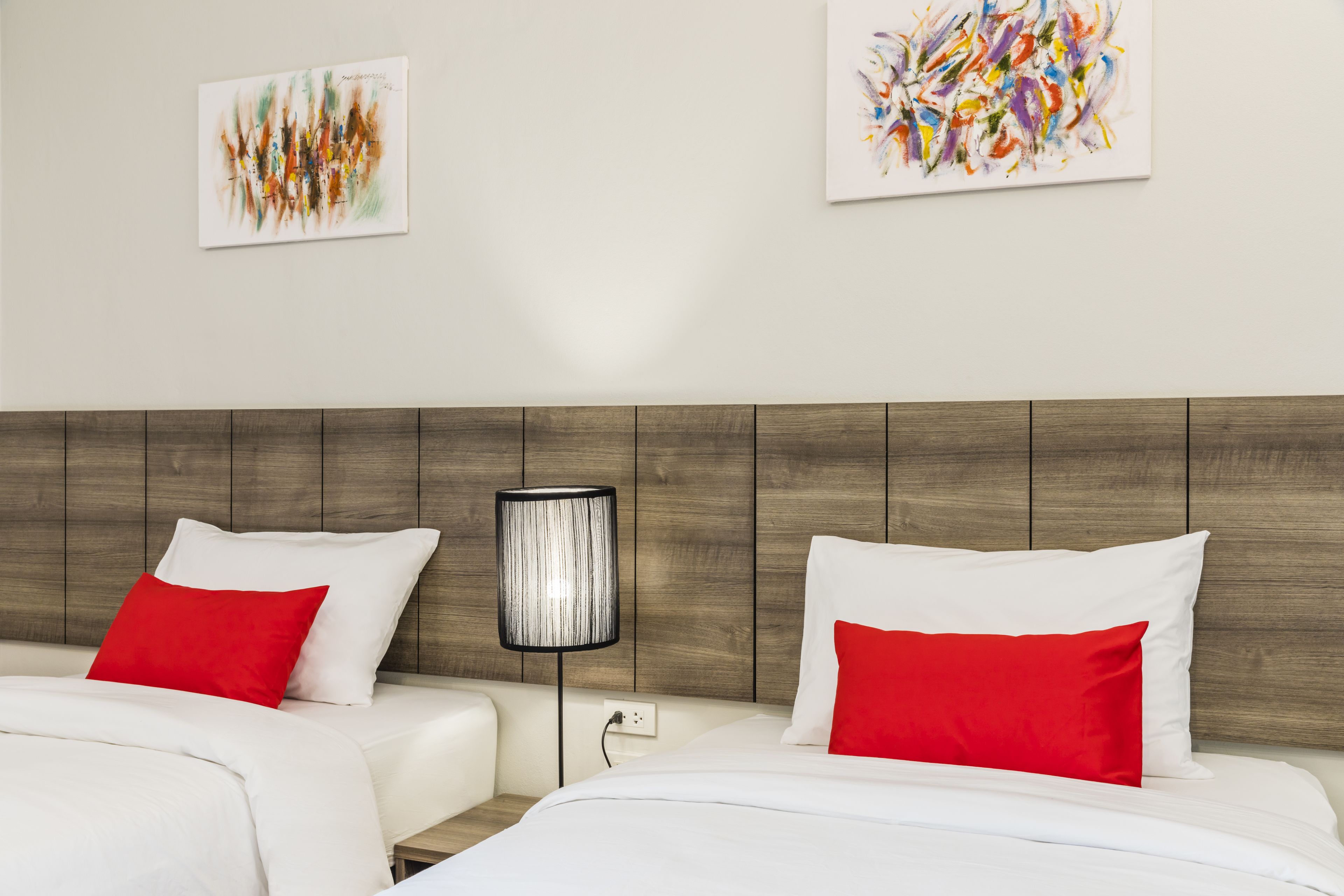 A hotel room with two beds, each with white bedding and red pillows, a wooden headboard, and abstract art on the walls.