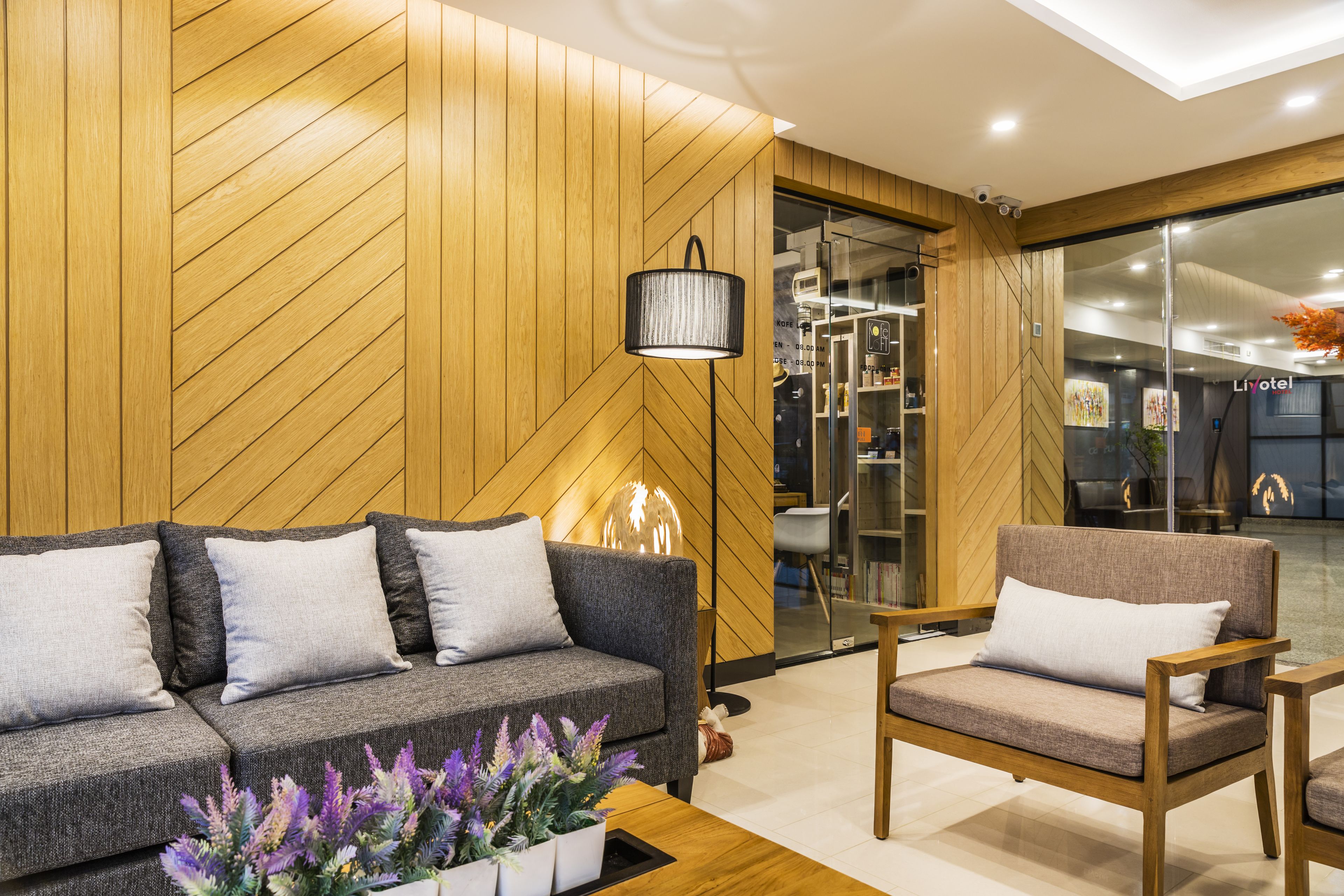 Un moderno vestíbulo de hotel con una pared de madera en patrón espiga, un sofá gris, un sillón de madera y una planta en maceta.