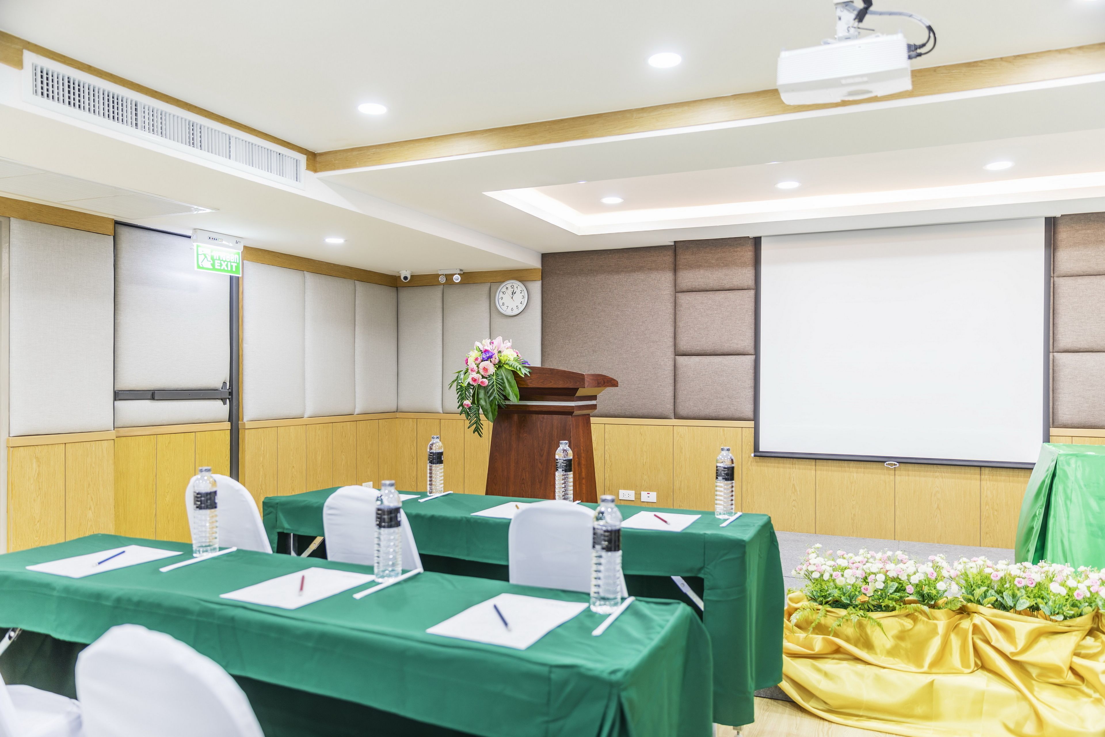 A conference room with tables, chairs, a podium, a projector, and an exit sign.
