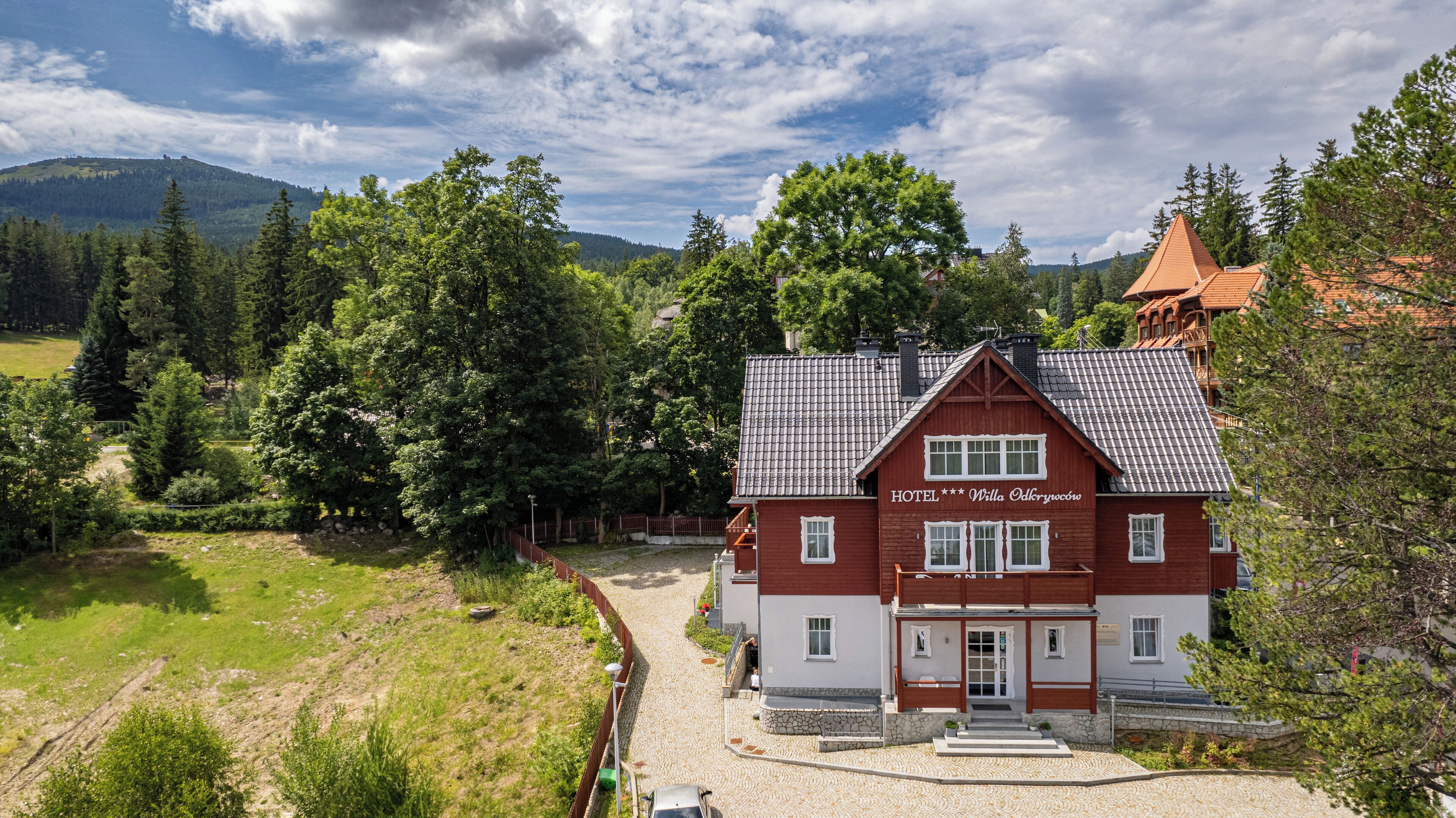 A red and white hotel with a tiled roof, surrounded by lush greenery and mountains.