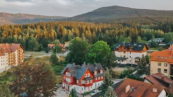 A residential area with a prominent red house with a sloped roof, surrounded by other buildings and a forested hillside in the background.
