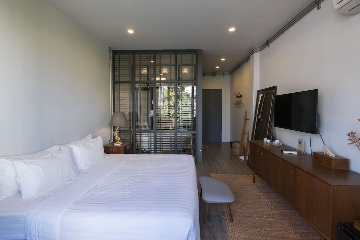 A modern hotel room with a large bed, a wooden dresser, a television, and a sliding glass door leading to a balcony.