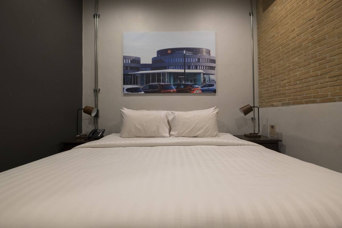 A hotel room with a large bed, two bedside tables, a wall-mounted telephone, and a framed picture of a building.