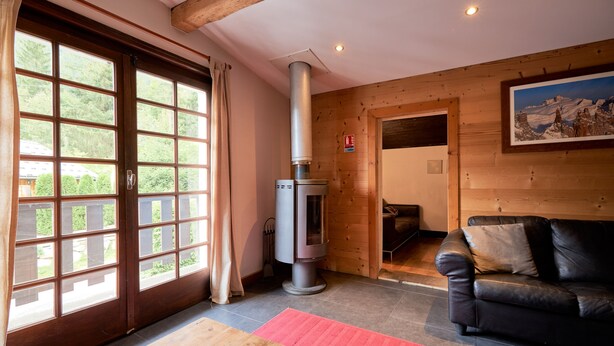 A cozy living room with a fireplace, a sofa, a wooden table, and large windows. - Chamonix Lodge (Chamonix-Mont-Blanc)