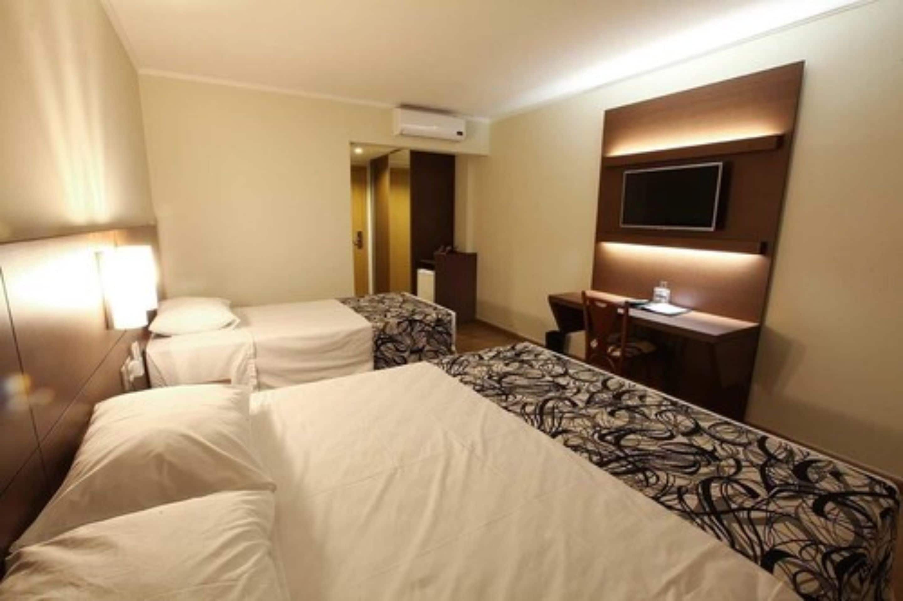 Une chambre d’hôtel avec deux lits simples, un bureau avec une chaise, un téléviseur à écran plat fixé au mur et un appareil de climatisation.