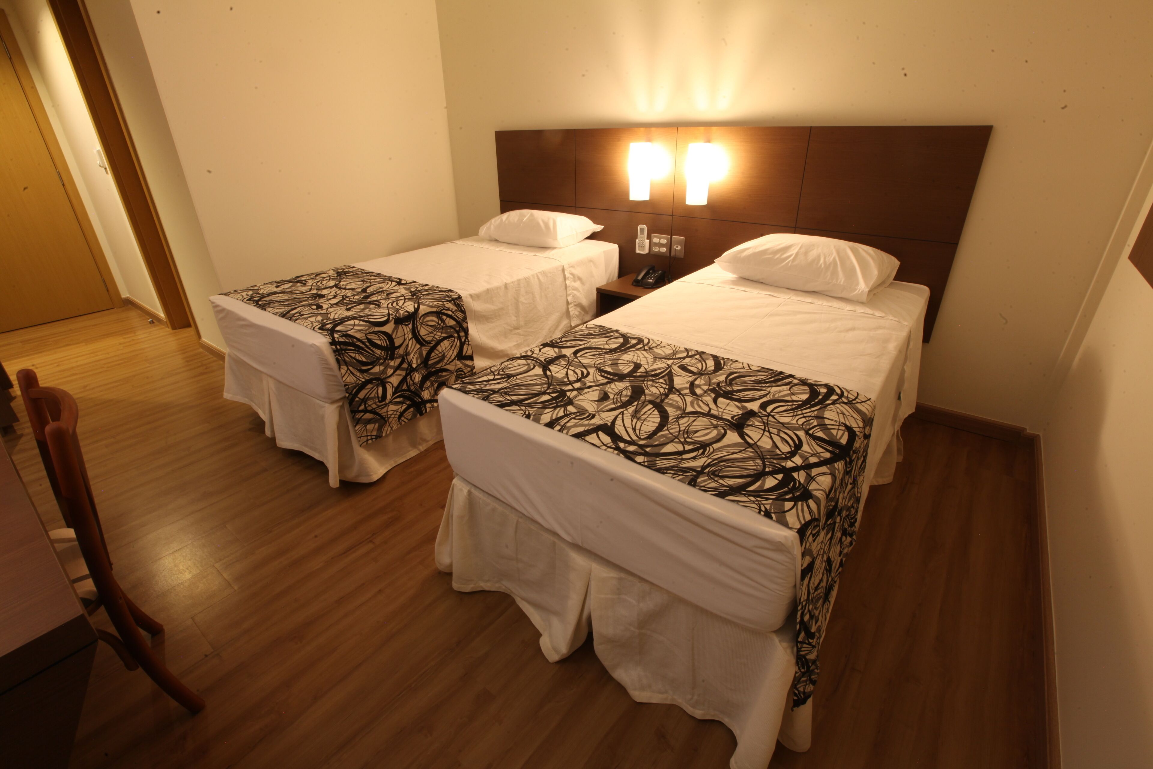 Habitación de hotel con dos camas, cada una con una colcha estampada y almohadas blancas. Hay piso de madera, una mesita de noche con teléfono y una lámpara de pared.