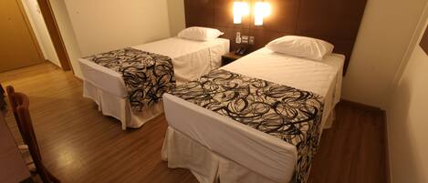 Habitación de hotel con dos camas, cada una con una colcha estampada y almohadas blancas. Hay piso de madera, una mesita de noche con teléfono y una lámpara de pared.