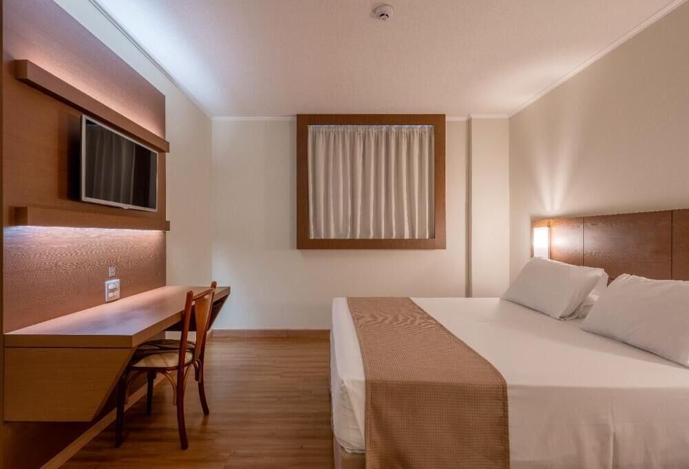 Ein Hotelzimmer mit einem Bett, einem Schreibtisch, einem Stuhl, einem an der Wand befestigten Fernseher und einem Fenster mit Vorhängen.