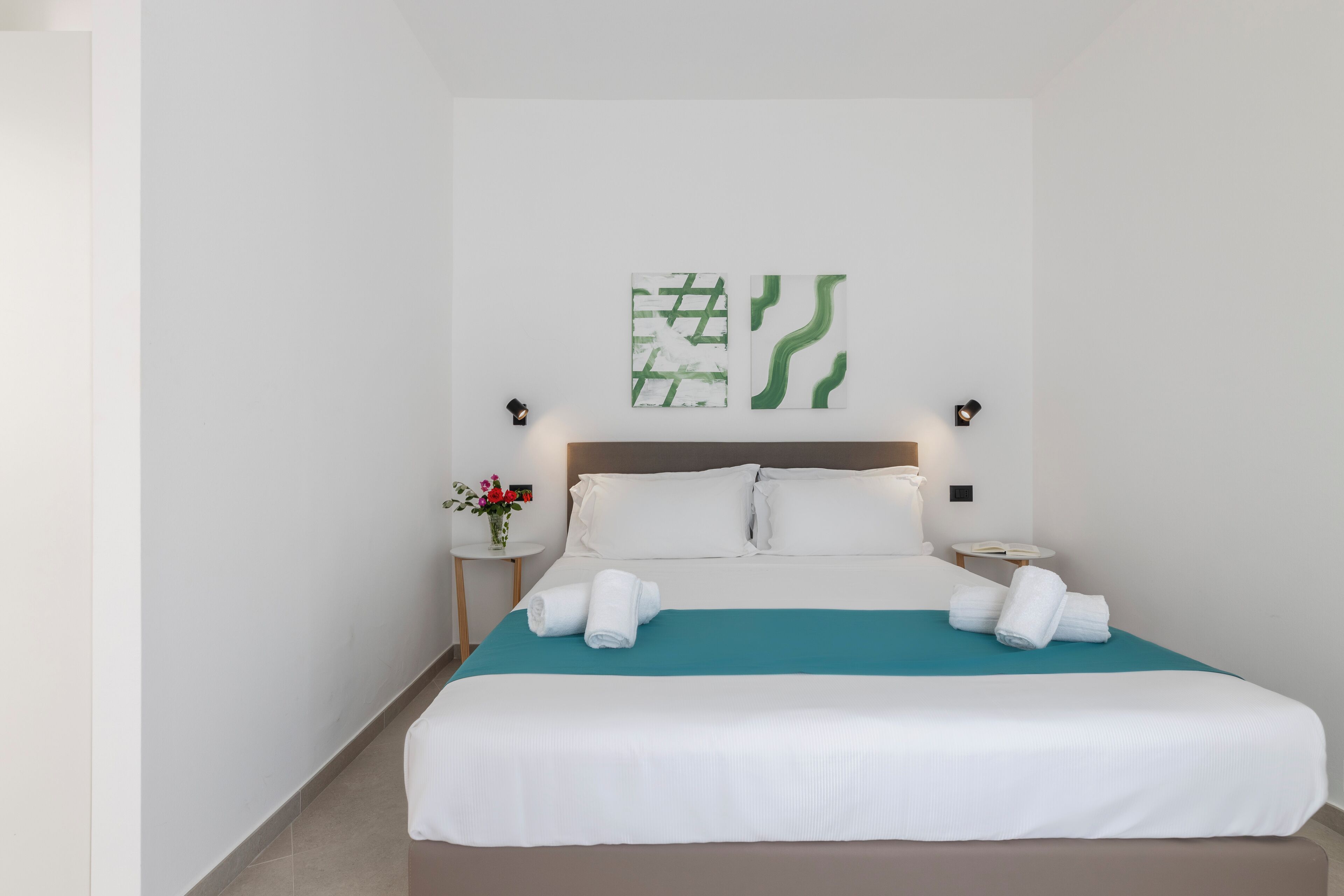 A hotel room with a bed, white bedding, a teal accent on the bedspread, two wall-mounted light fixtures, and two abstract green and white wall art pieces.