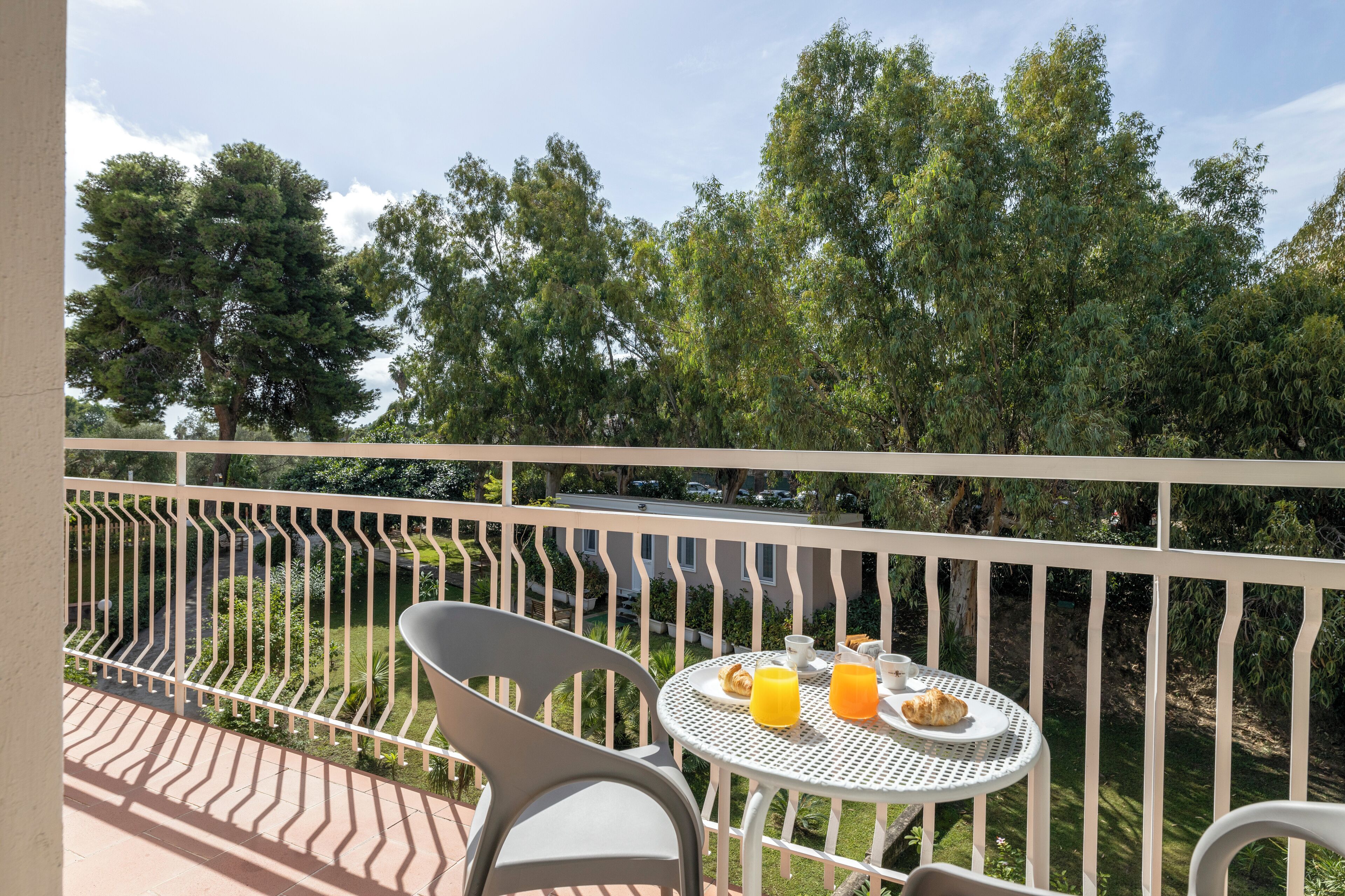 Ein Balkon mit einem weißen Metalltisch und Stühlen, einem Teller mit Croissants und Orangensaft sowie Blick auf Bäume und eine Straße.