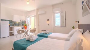 A hotel room with two beds, a kitchenette, a dining area, and a large window. - Residence Villa Giardini (Giardini Naxos)